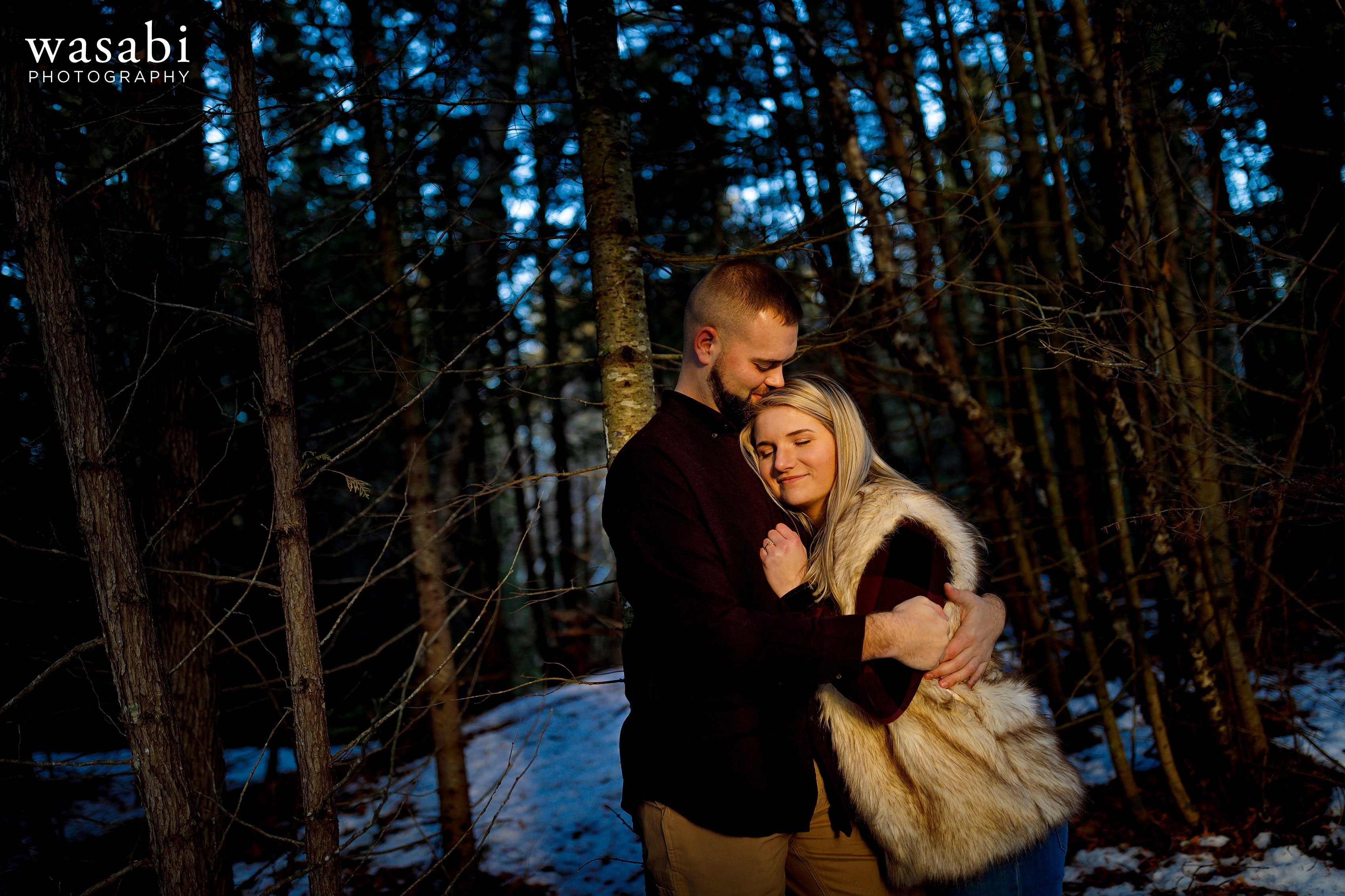 couple poses for engagement photo with coniferous trees in the background during winter engagement session in Glen Arbor Michigan