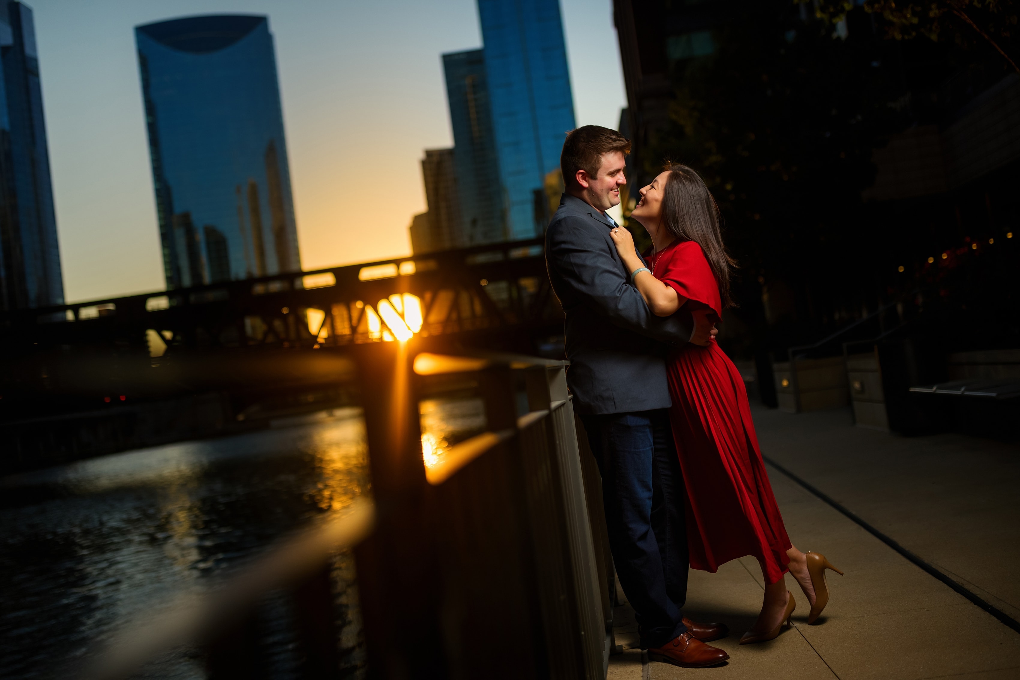 Couple poses for engagement photos along riverwalk with the sun setting in the background in downtown Chicago