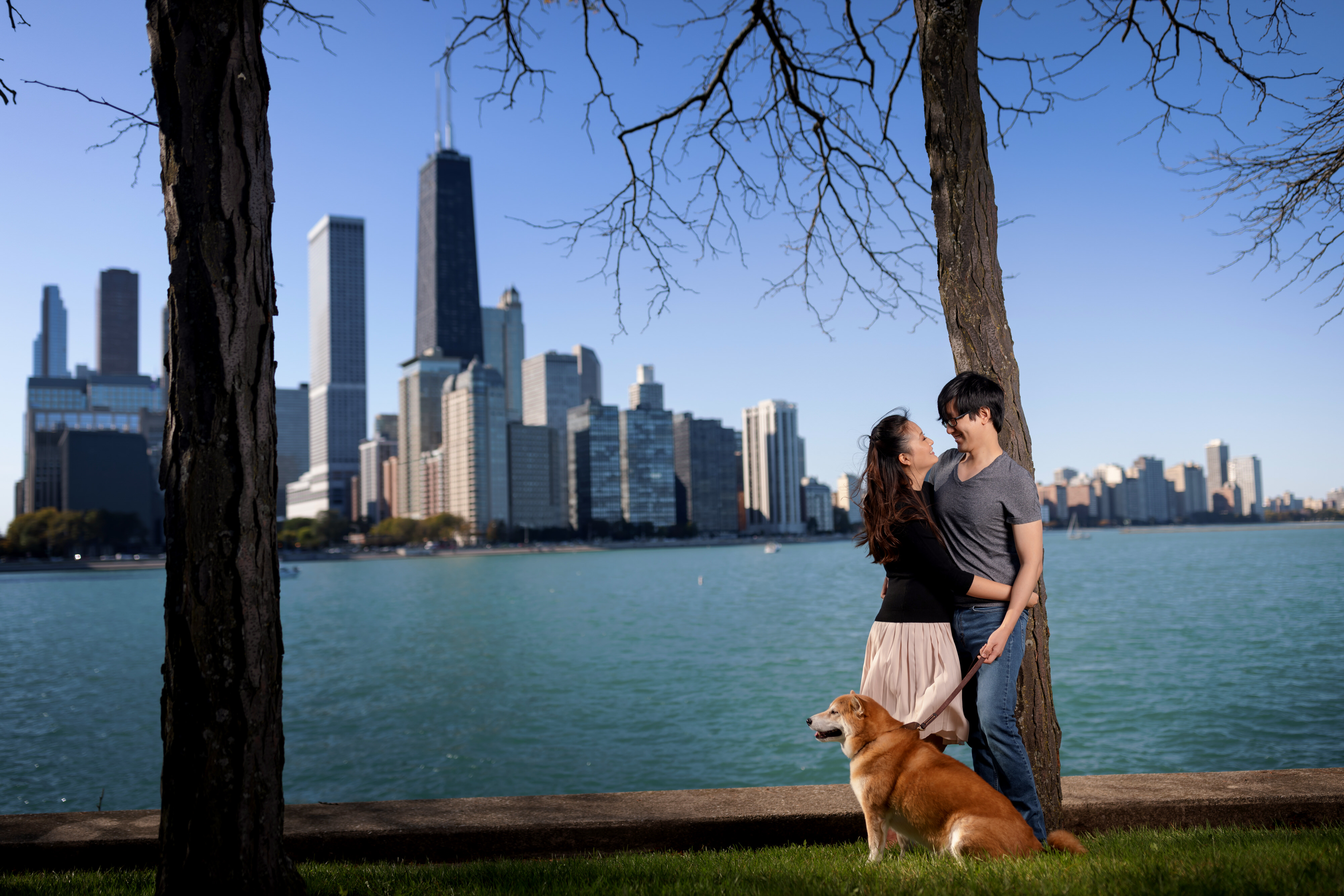 couple poses for engagement photos with their dog at milton lee olive park in chicago with lake michigan and the skyline in the background