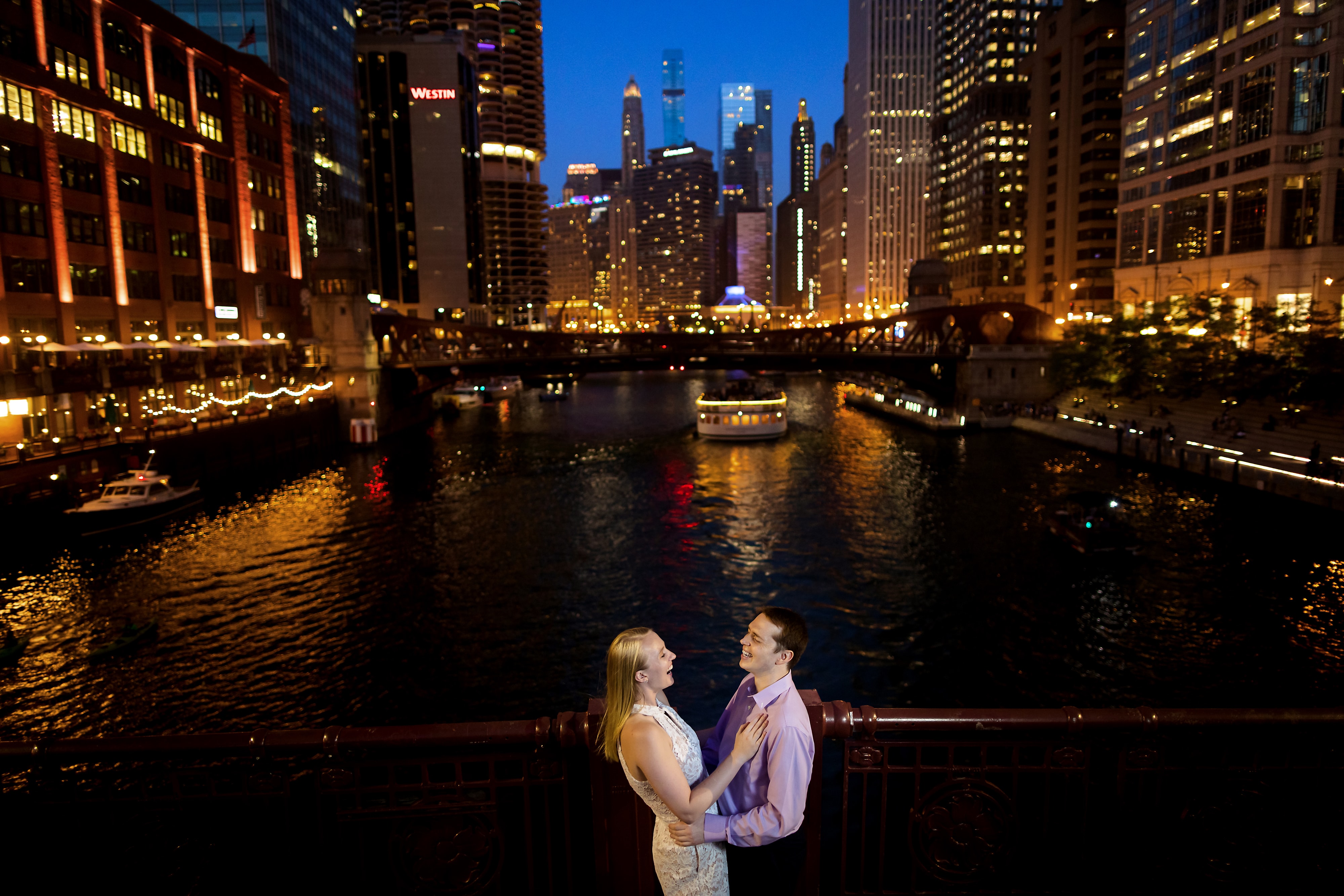 couple laughs with the Chicago river and skyline in the background on LaSalle Street Bridge during summer engagement session at night in downtown Chicago