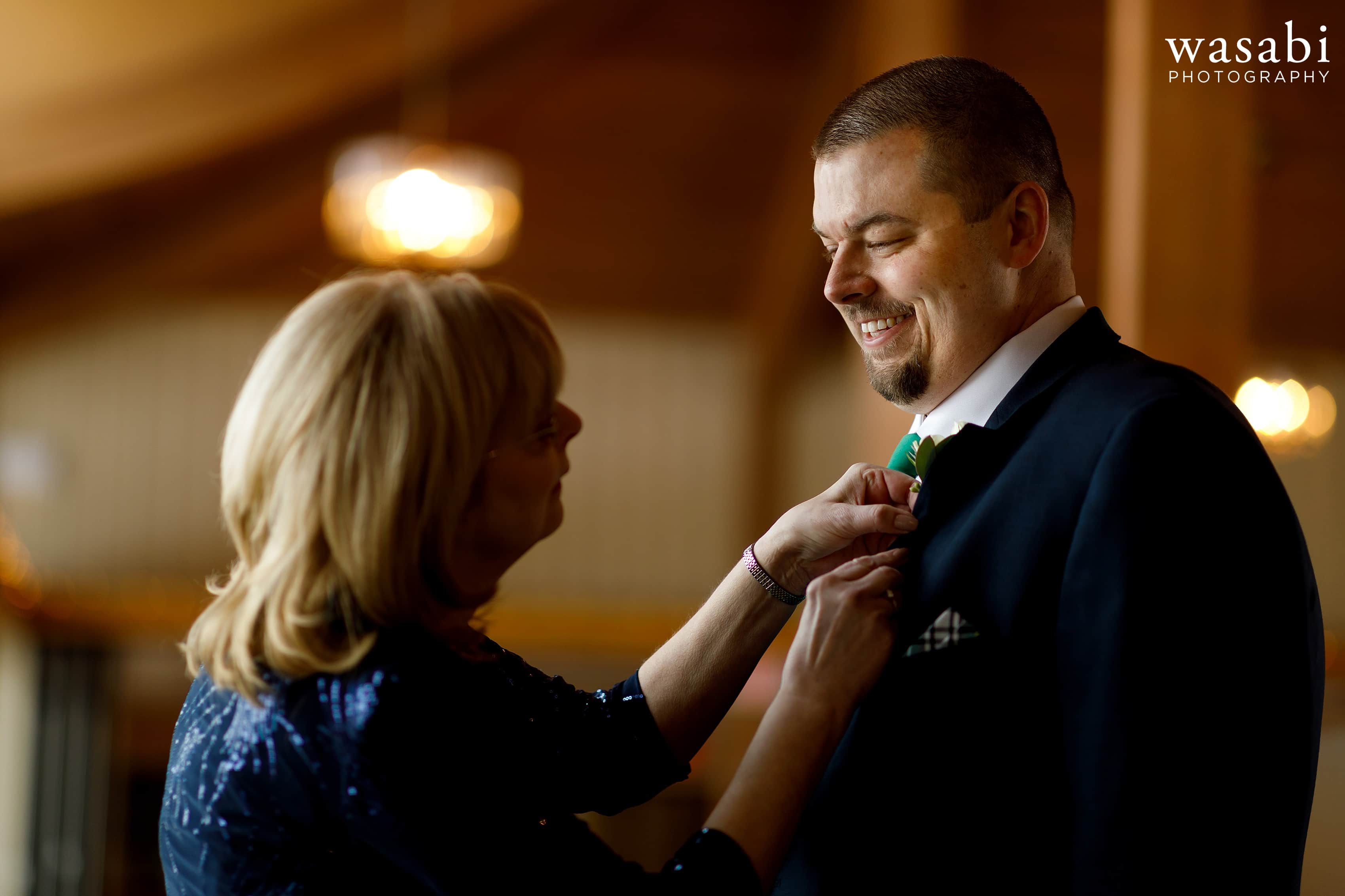 mother of groom puts boutonnière on son's jacket while getting ready for wedding