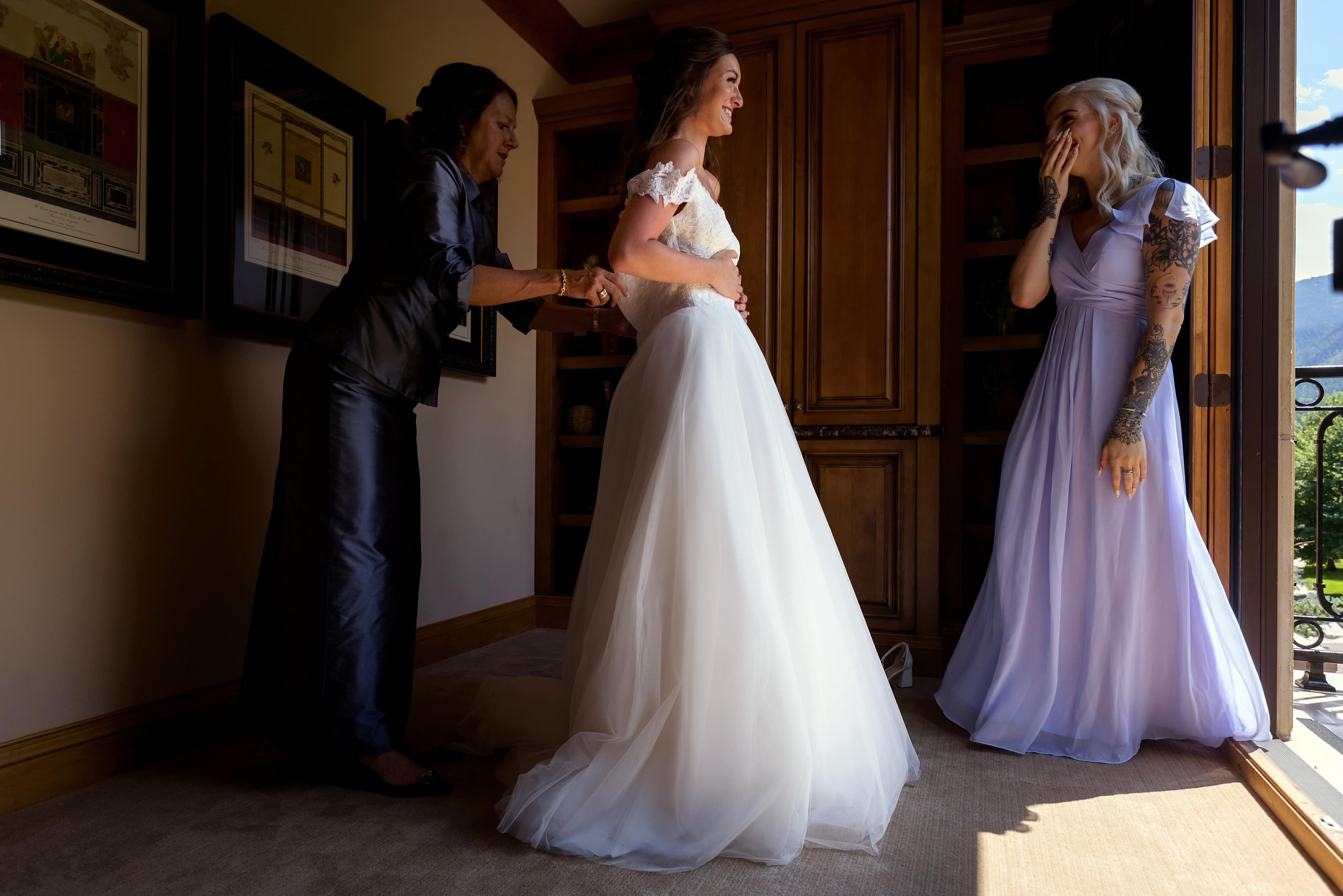 mother of bride and sister help bride into dress while getting ready