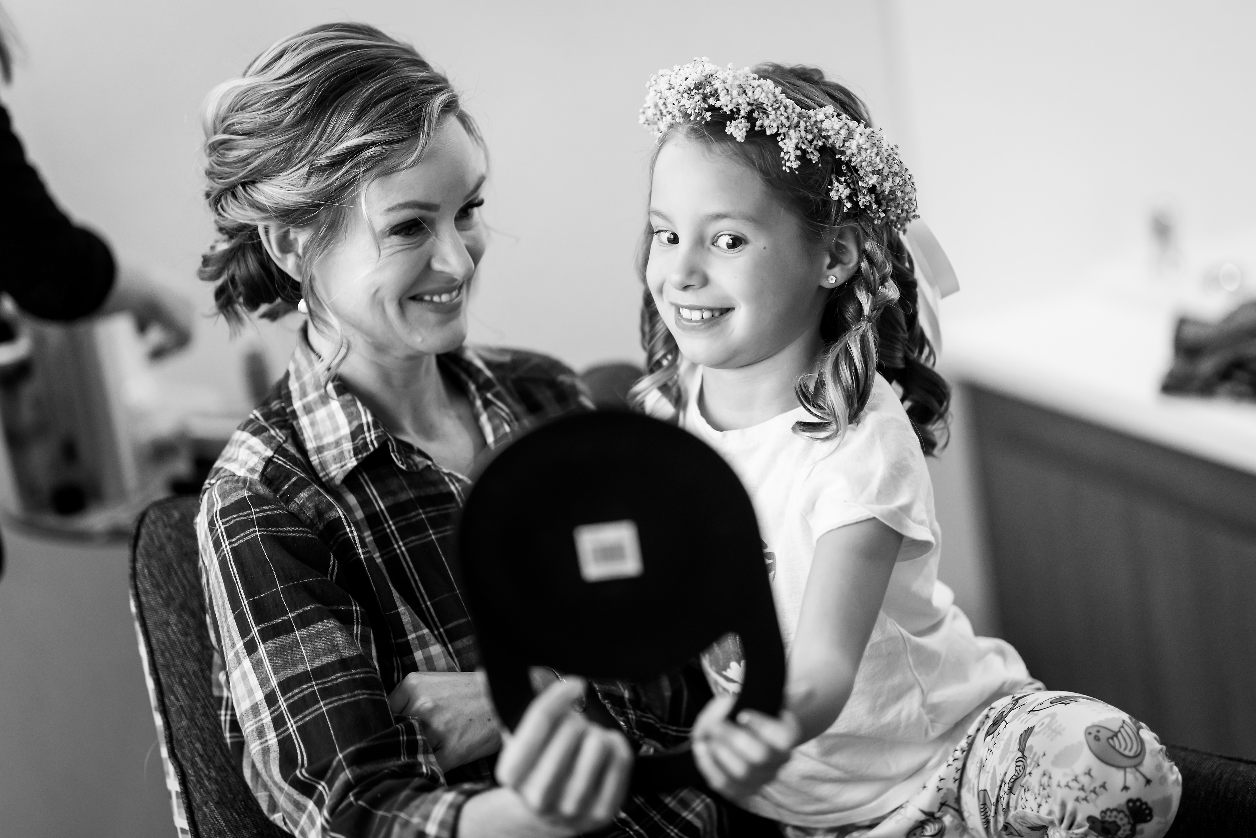 Mom and flower girl daughter look into mirror while getting ready for wedding