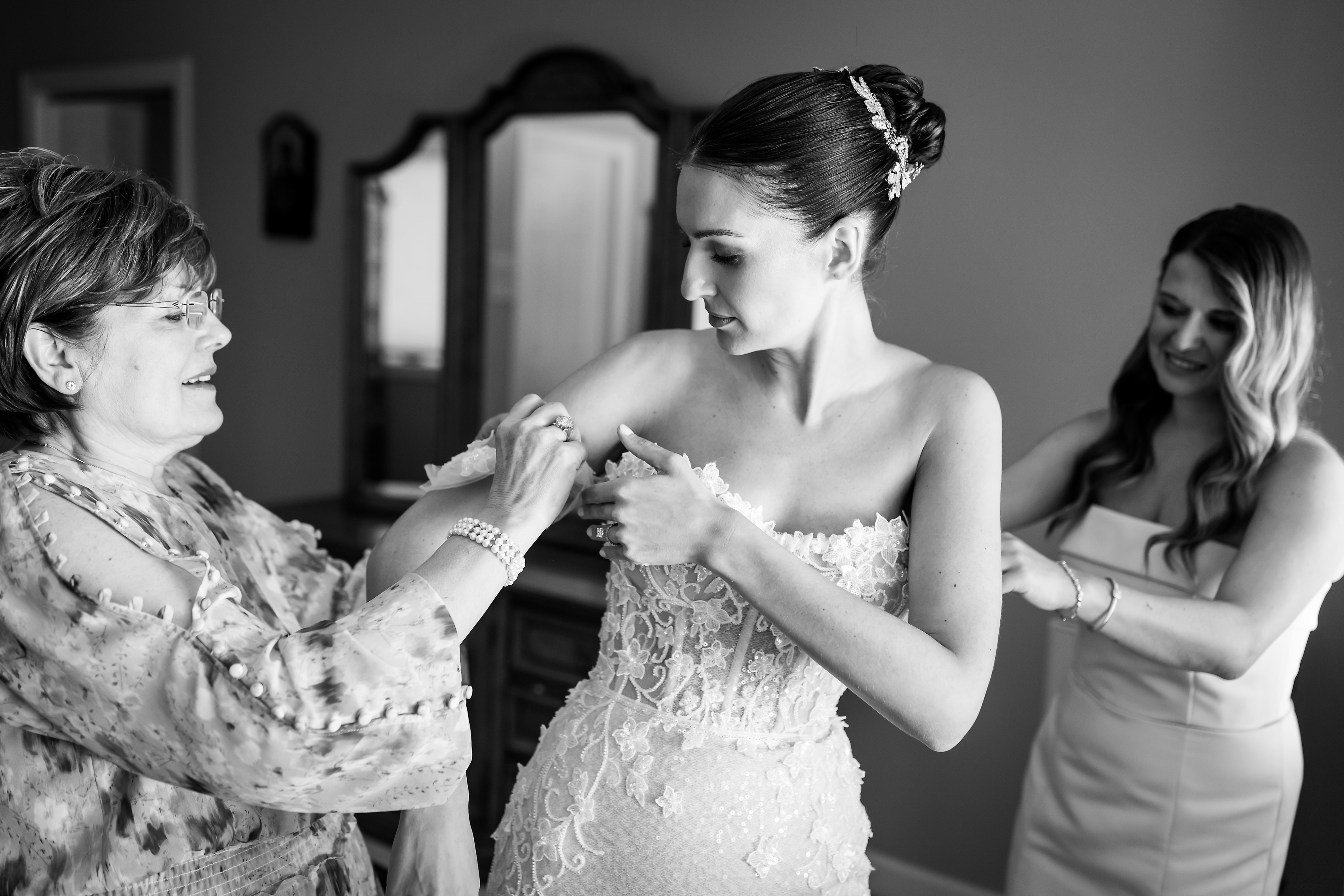 Mother and bridesmaid help bride put on wedding dress