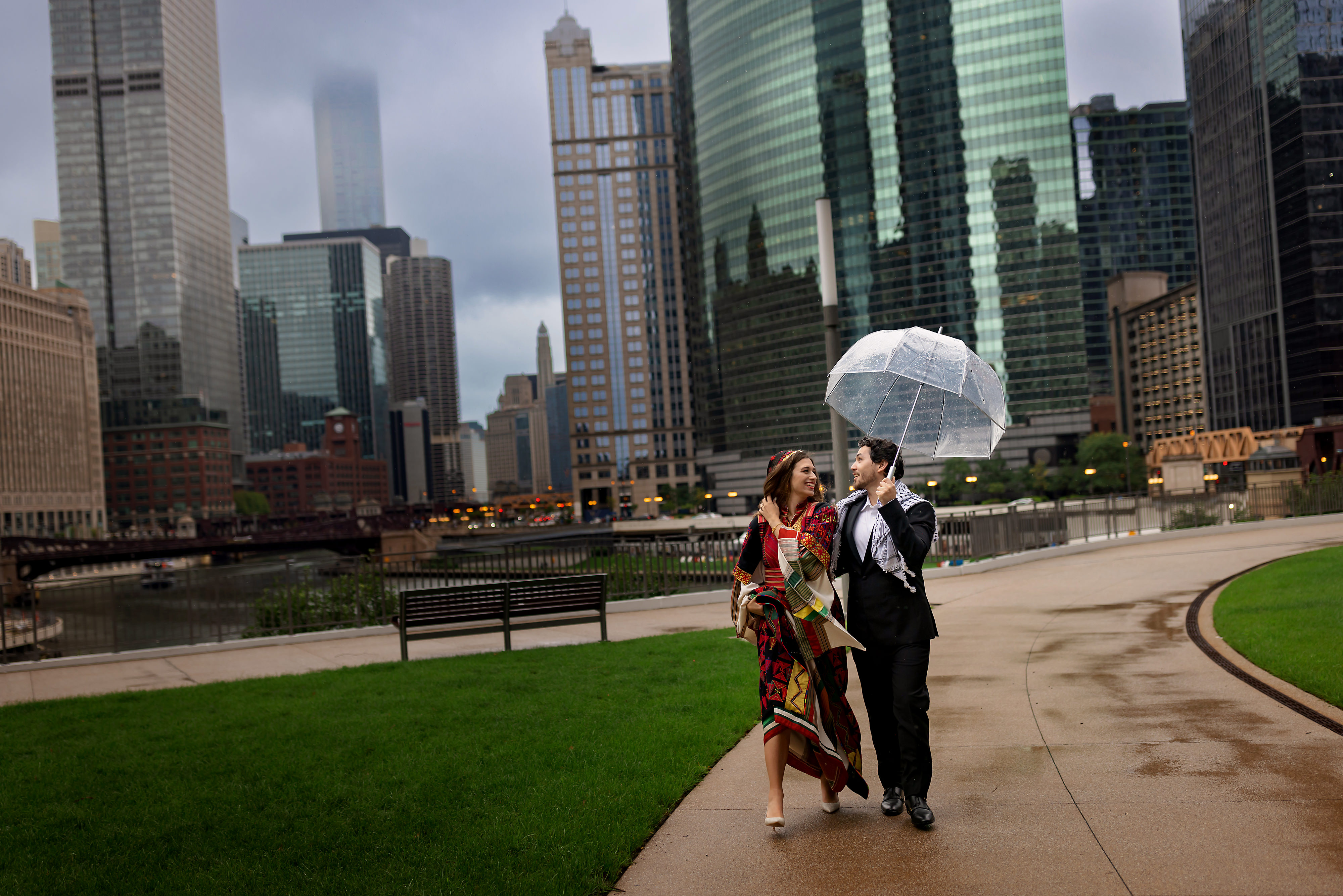 Bride and groom walk under umbrella near Gibson's Italia Plaza Riverwalk with the Chicago skyline and river in the background