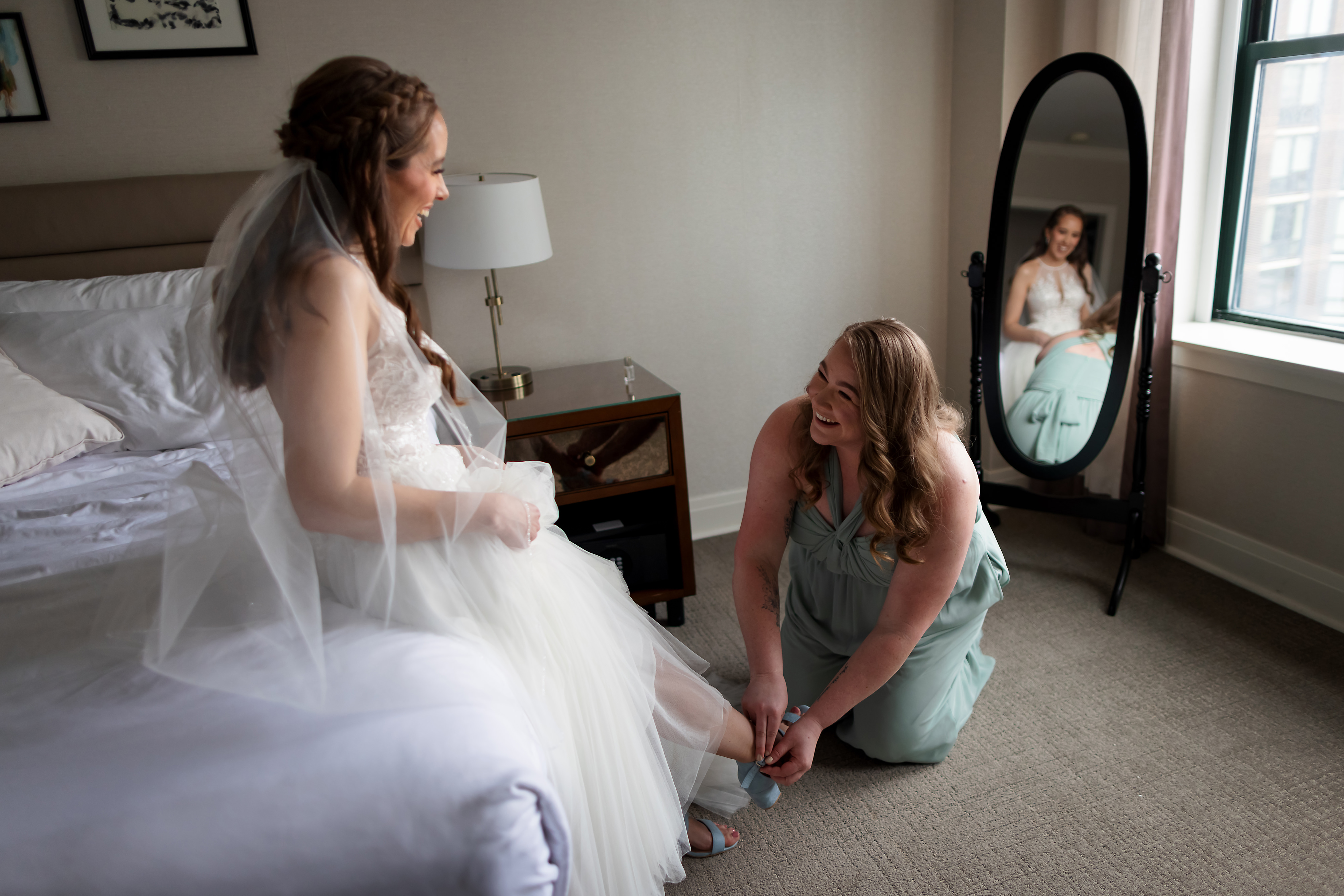 Bridesmaid puts shoe on bride while looking into mirror while getting ready for wedding in Lincoln Park, Chicago