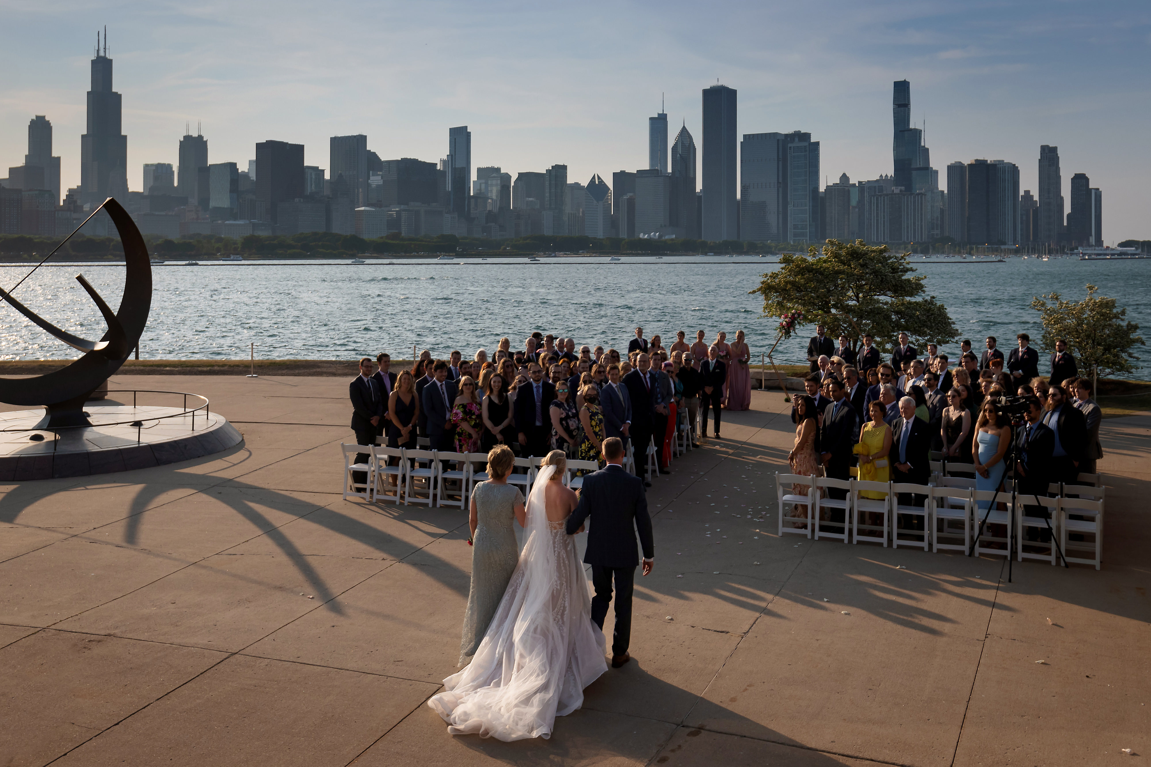 wide angle view of bride walking down the aisle with parents and Chicago city skyline in the background during wedding ceremony at Adler Planetarium in Chicago